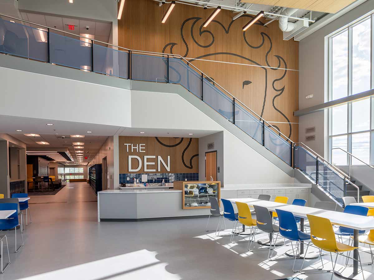 Modern school commons area with tables, blue and yellow chairs, a staircase, and a large lion mural labeled “The Den.”